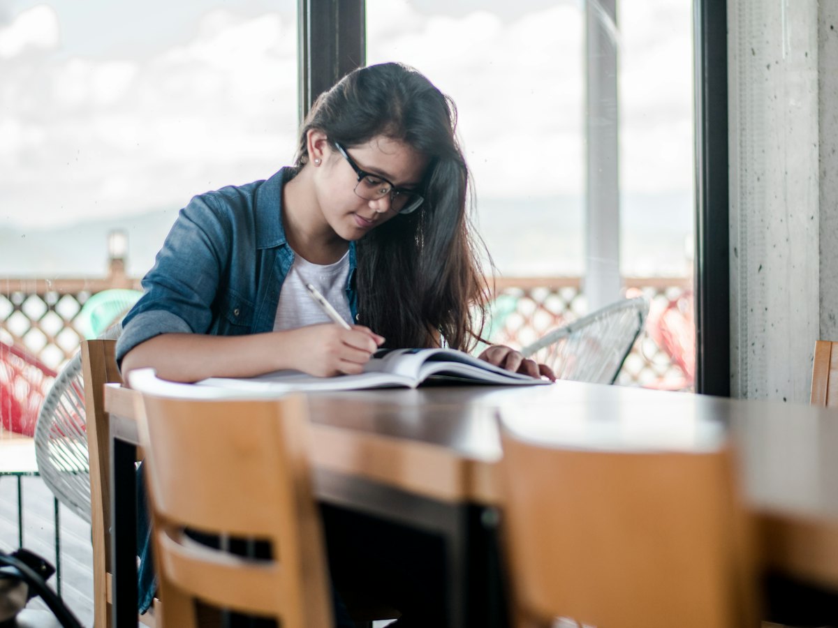 A student writing in a notebook at a table by the window.