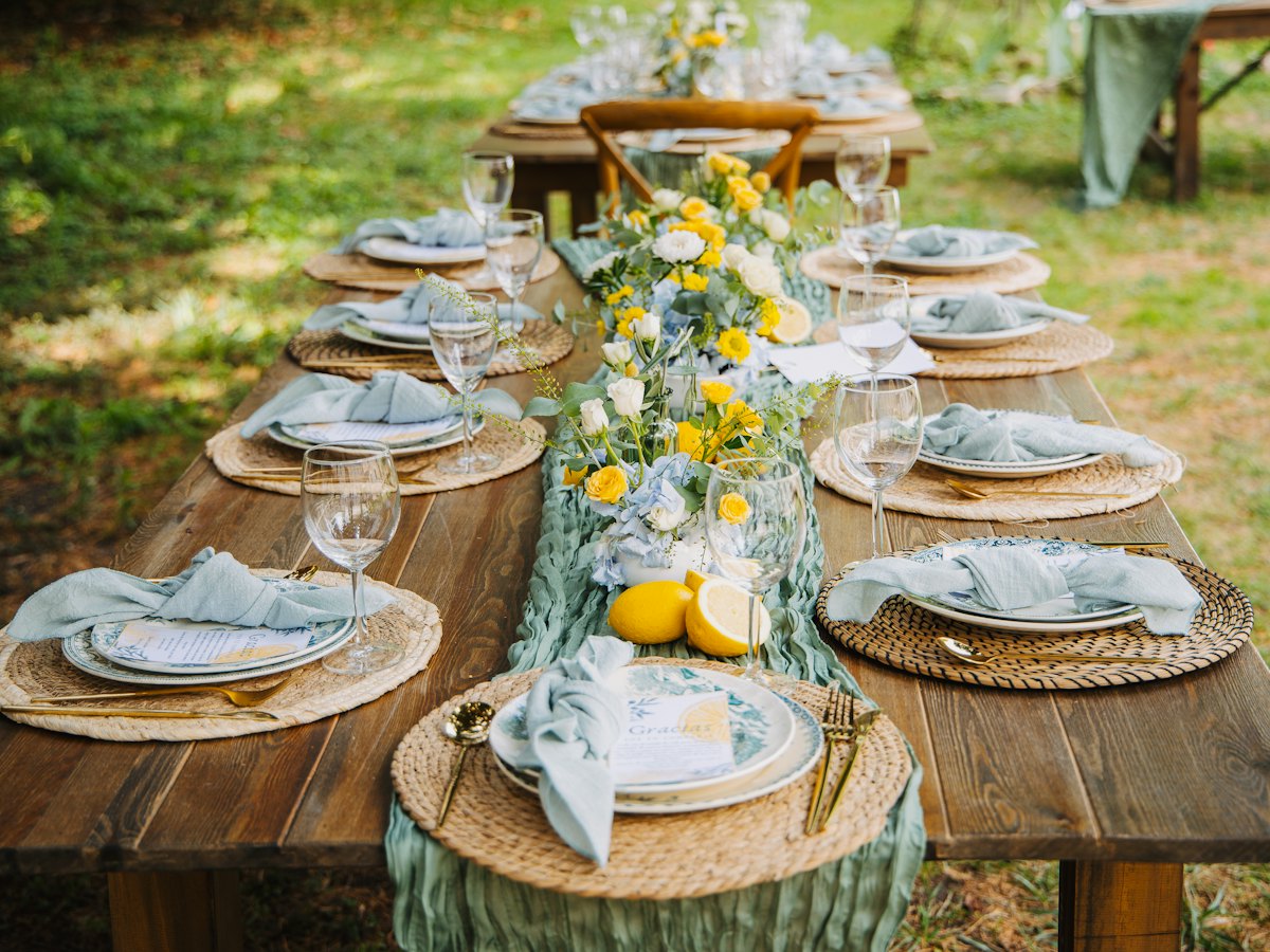 A long wooden table set for dinner outdoors, with lemons and greenery.